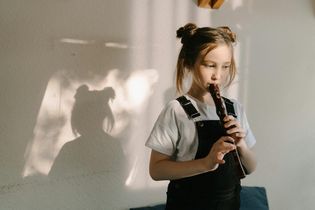 A young girl plays a flute indoors with sunlight casting a shadow on the wall.